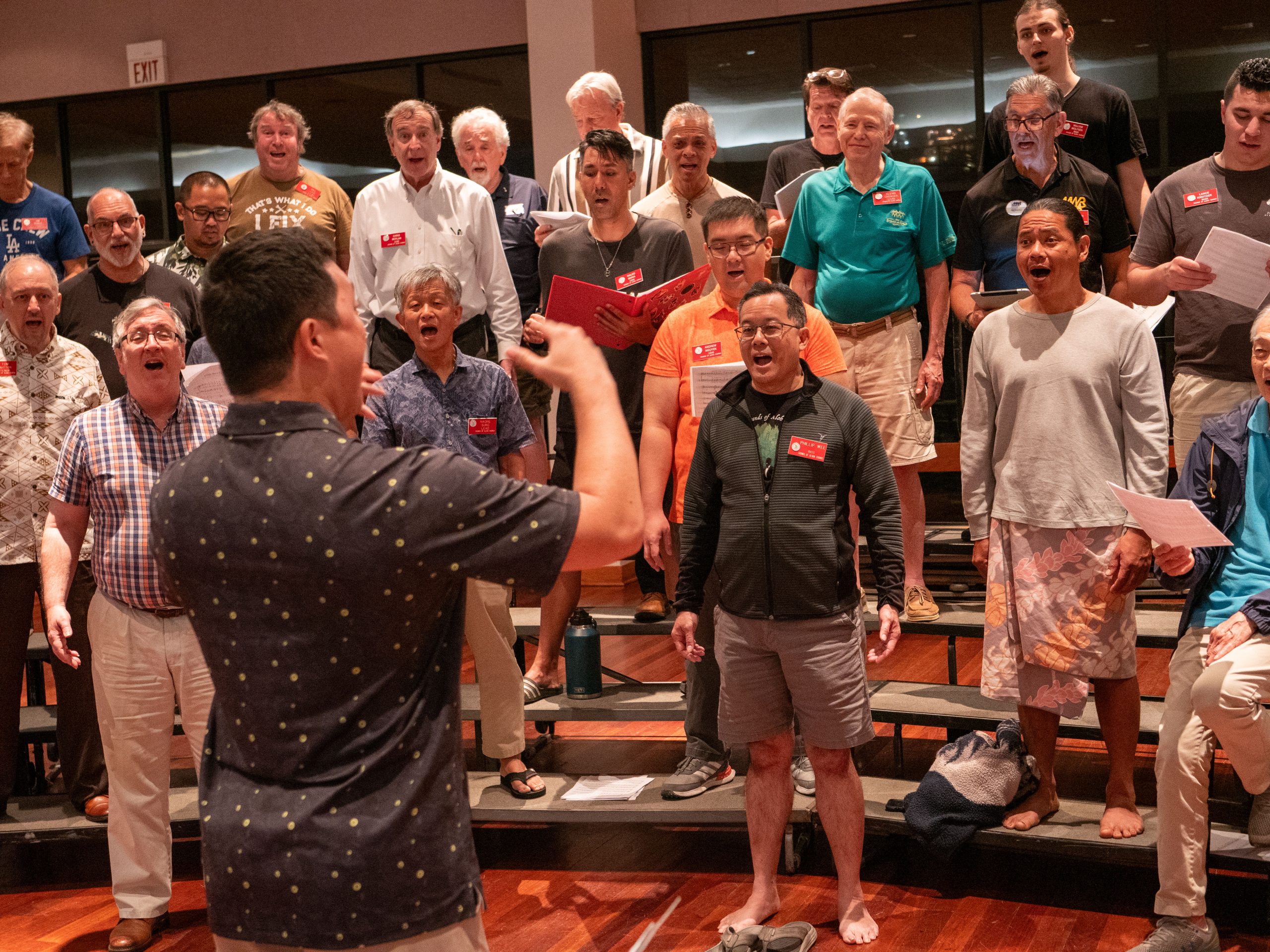 A large group of men standing on tiered steps, singing together as a choir, while a conductor facing them raises his arms in front of the group.